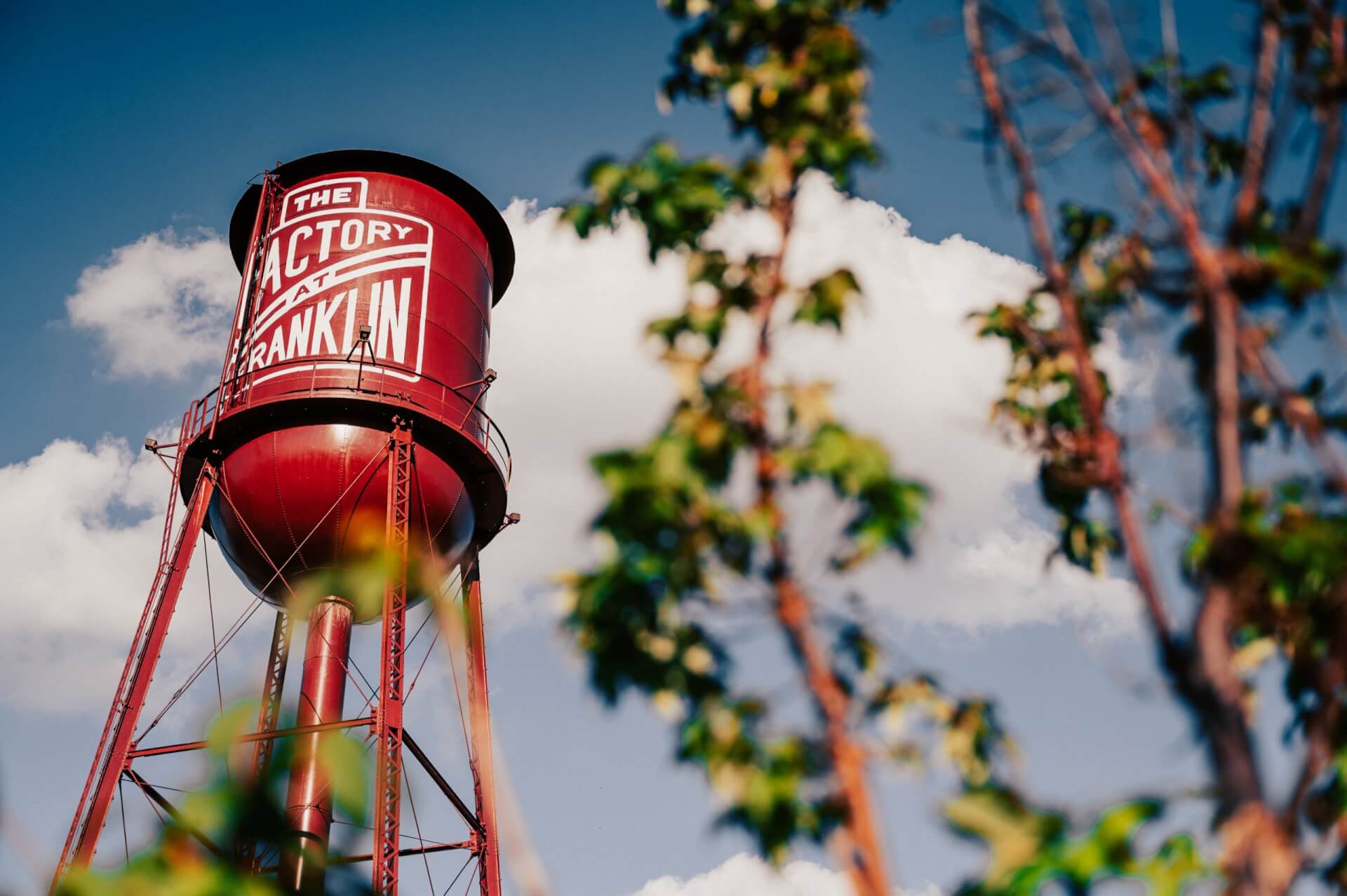 A tall, red water tower with white lettering stands against a backdrop of a clear blue sky with fluffy white clouds in Williamson County, Tennessee.