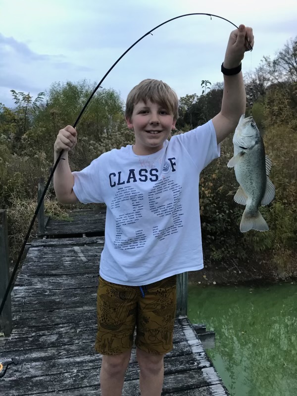 A young boy proudly displays a freshly caught fish on a wooden dock, wearing a 'Class of 2024' t-shirt and patterned shorts in Williamson County, Tennessee.