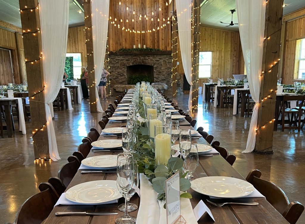 Elegant wedding reception venue with long wooden tables adorned with white plates, green garlands, and tall candles, set in a rustic barn with string lights and white curtains in Williamson County, Tennessee.