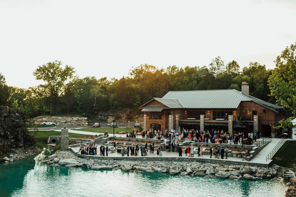 A large group of people gathers on a stone patio beside a serene lake, with a rustic wooden building and lush greenery in the background in Williamson County, Tennessee.