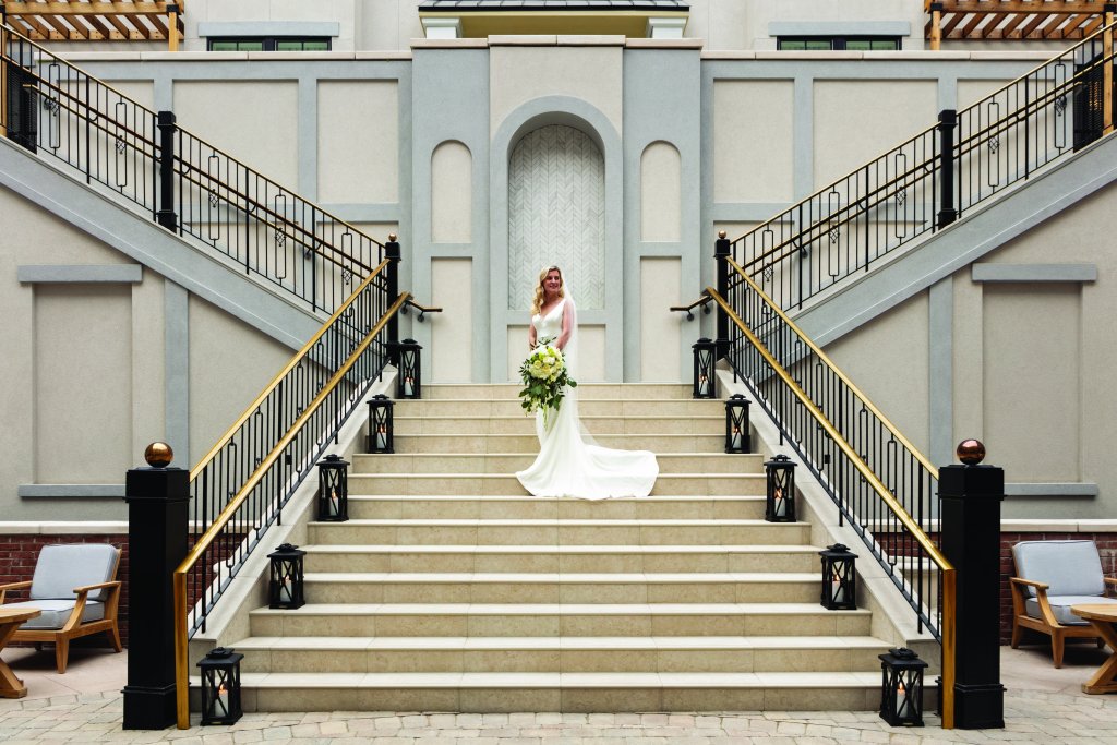 A bride in a flowing white gown stands at the top of an elegant staircase, holding a bouquet of white flowers, with a grand architectural backdrop in Williamson County, Tennessee.
