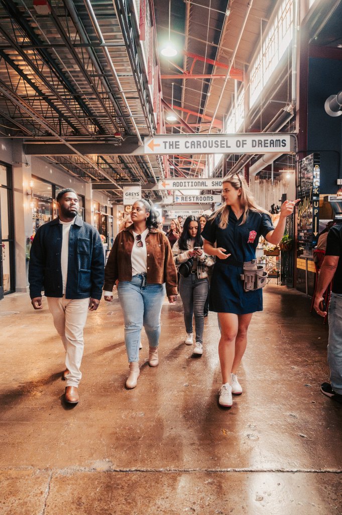 A group of people stroll through a bustling indoor market with high ceilings and exposed beams, illuminated by natural light streaming through large windows in Williamson County, Tennessee.