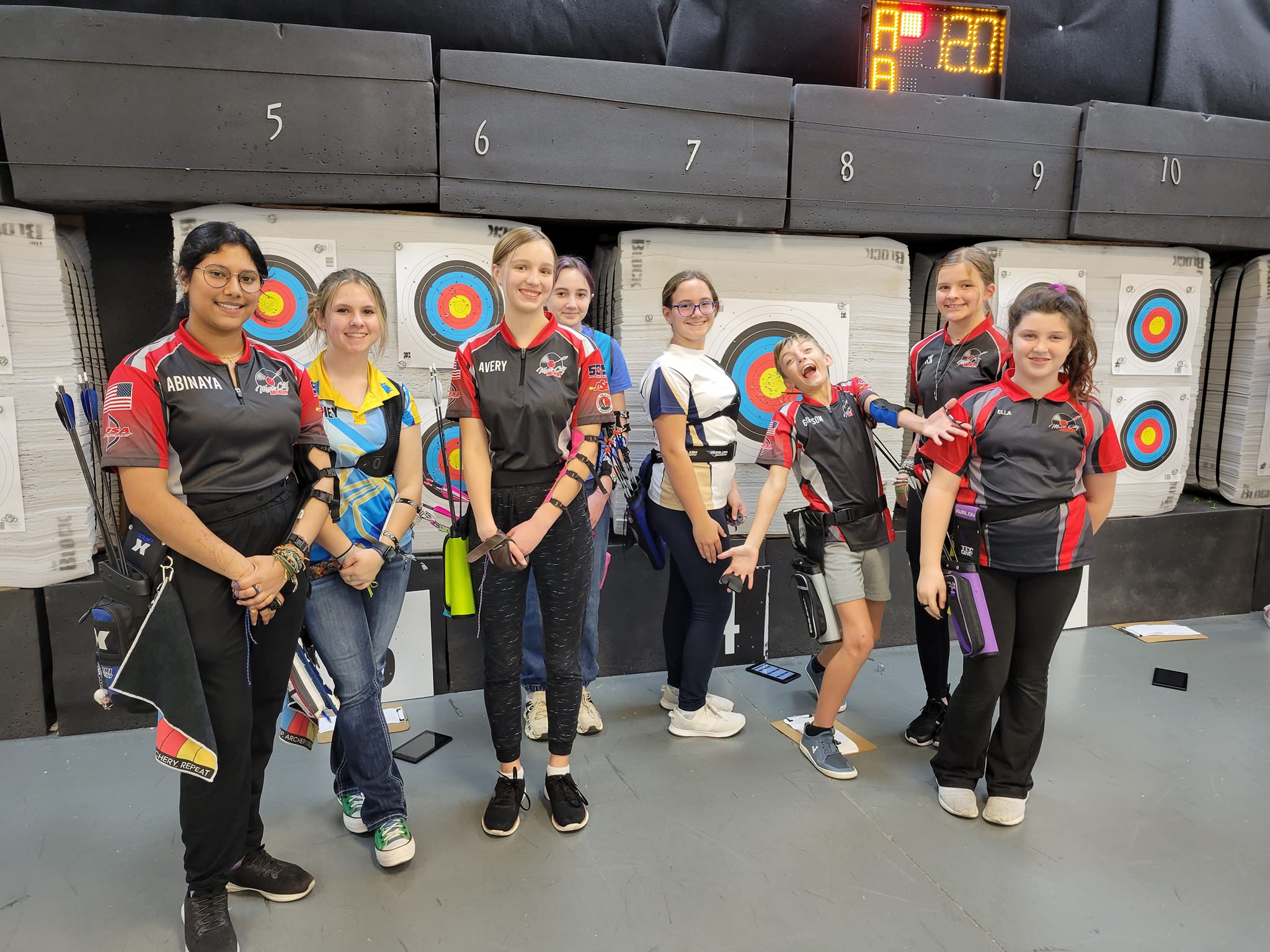 A group of eight young women, dressed in matching archery uniforms, pose together in front of a wall of archery targets in an indoor range in Williamson County, Tennessee.