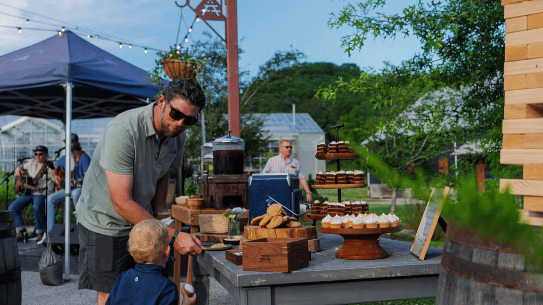 A man and child at an outdoor market table filled with baked goods and produce under a blue tent with string lights in Williamson County, Tennessee.