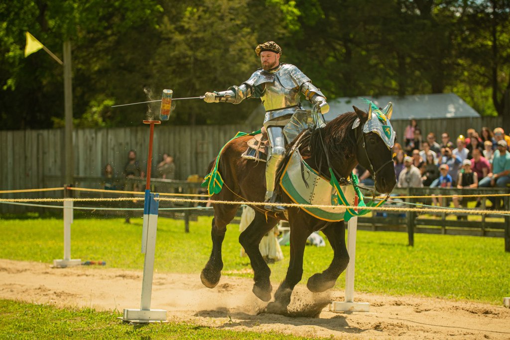 Tennessee Renaissance Festival A knight in full armor charges on horseback, brandishing a lance at a target, while spectators watch from behind a fence in Williamson County, Tennessee.