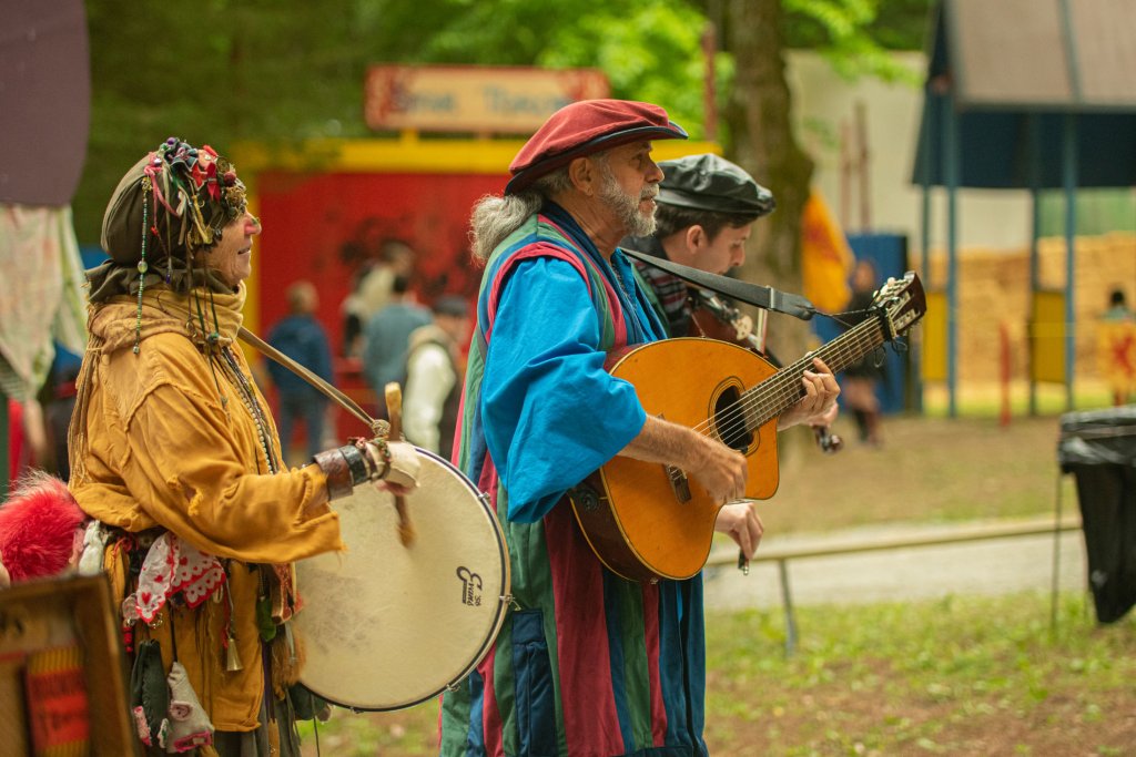 Tennessee Renaissance Festival Two musicians dressed in colorful, traditional costumes play a drum and guitar in an outdoor setting, surrounded by a crowd and a festive atmosphere in Williamson County, Tennessee.