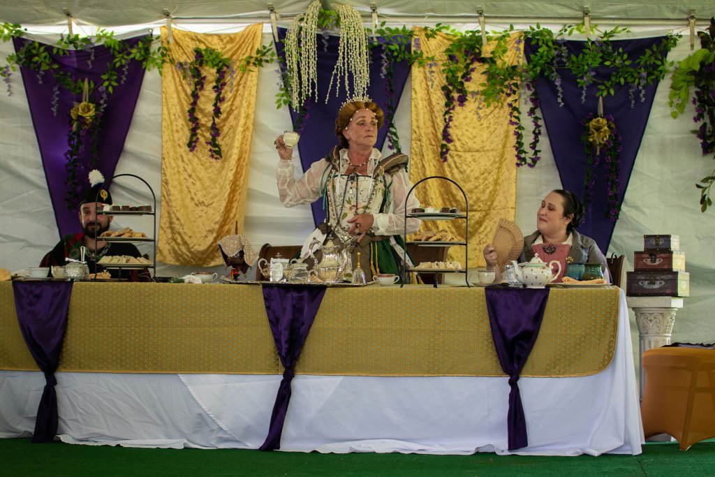 Tennessee Renaissance Festival A woman dressed in a regal costume stands at a lavishly decorated table, holding a teacup, with two other costumed individuals seated beside her in Williamson County, Tennessee.