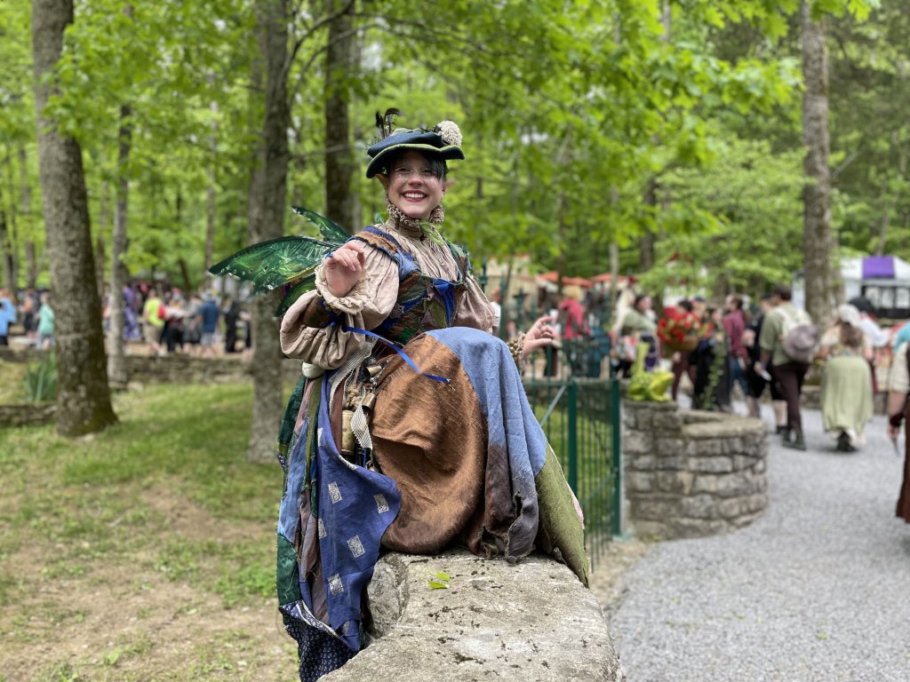 Tennessee Renaissance Festival A woman dressed in a whimsical fairy costume with green wings and a hat sits smiling on a stone wall in a lush forest setting, surrounded by a crowd of people in period attire in Williamson County, Tennessee.