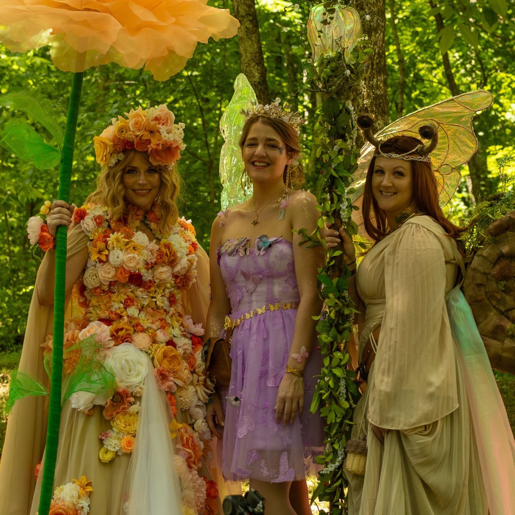 Tennessee Renaissance Festival Three women dressed as fairies, adorned with vibrant floral crowns and wings, stand together in a lush forest, smiling and holding a large orange flower and a staff with glowing orbs in Williamson County, Tennessee.