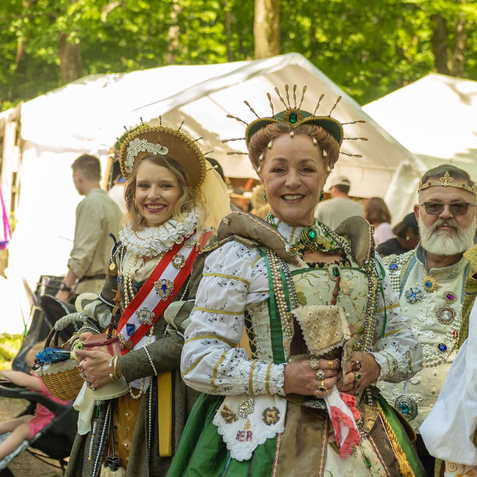 Tennessee Renaissance Festival Two women dressed in elaborate Renaissance-era costumes, adorned with jewels and crowns, stand smiling amidst a festive outdoor gathering with white tents and trees in the background in Williamson County, Tennessee.