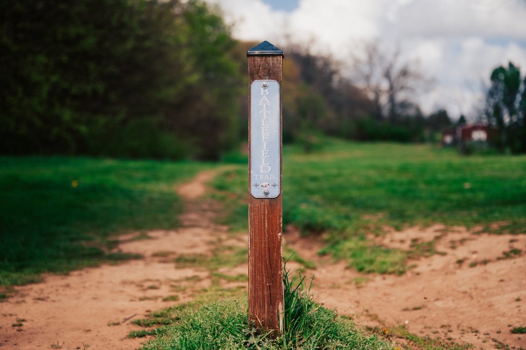 A wooden post with a sign reading 'Battlefield Trail' stands at the start of a dirt path in a grassy field, surrounded by trees in Williamson County, Tennessee.