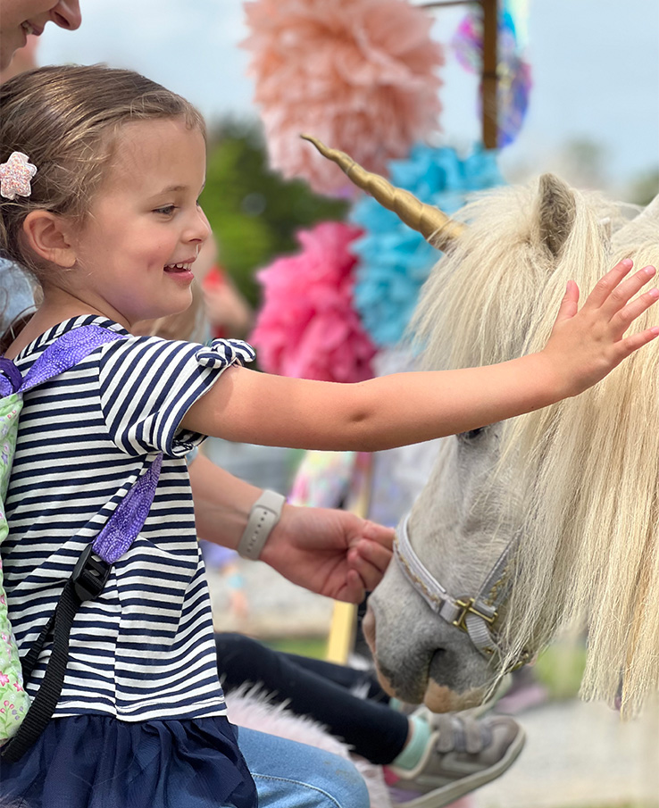 A young girl with a striped shirt and a unicorn horn headband gently pets a white miniature horse with a golden horn at a whimsical outdoor event in Williamson County, Tennessee.