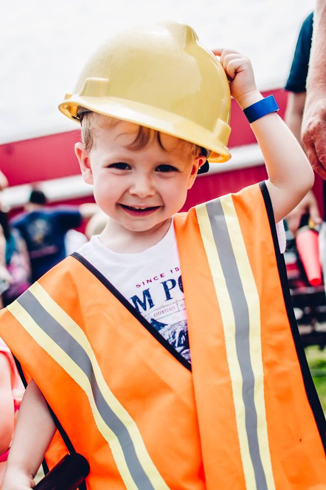 A young child dressed in a construction worker costume, complete with a yellow hard hat and an orange safety vest with reflective stripes, smiles brightly while holding the hat on their head in Williamson County, Tennessee.