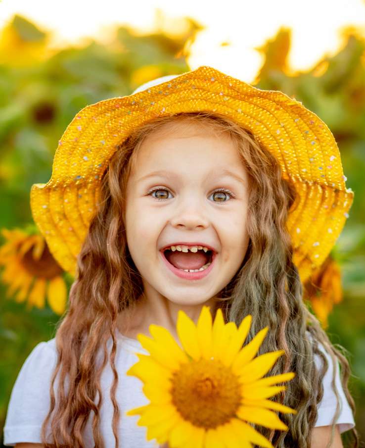 A young girl with long, curly hair is joyfully holding a vibrant sunflower, wearing a yellow hat adorned with colorful sprinkles, and standing amidst a field of sunflowers bathed in warm sunlight in Williamson County, Tennessee.