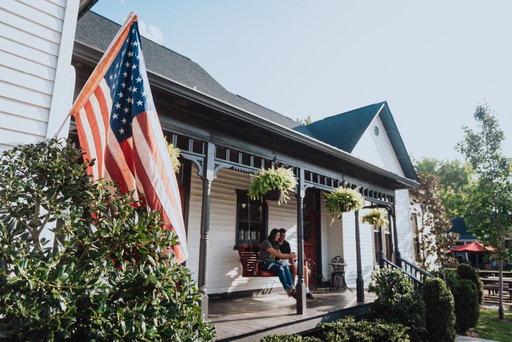 Plan the Perfect Reading Retreat in Franklin, TN with These Cozy Bookstores & Scenic Nooks A couple relaxes on a porch swing under a large American flag, enjoying a sunny day at a quaint white house with black trim in Williamson County, Tennessee.