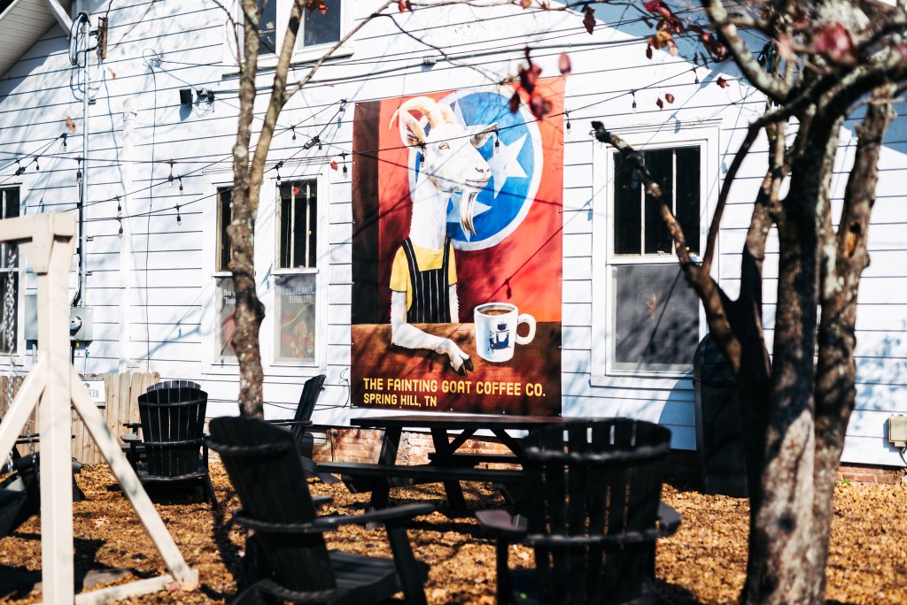 Plan the Perfect Reading Retreat in Franklin, TN with These Cozy Bookstores & Scenic Nooks A whimsical mural of a goat wearing overalls and holding a coffee mug adorns the side of a white building, surrounded by outdoor seating and autumn leaves in Williamson County, Tennessee.