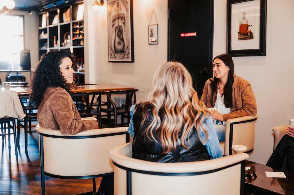 Plan the Perfect Reading Retreat in Franklin, TN with These Cozy Bookstores & Scenic Nooks Three women are engaged in a lively conversation in a cozy café setting, surrounded by comfortable seating and framed artwork in Williamson County, Tennessee.