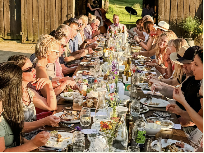 A large group of people enjoying a communal meal at a long table set outdoors under a wooden structure in Williamson County, Tennessee.