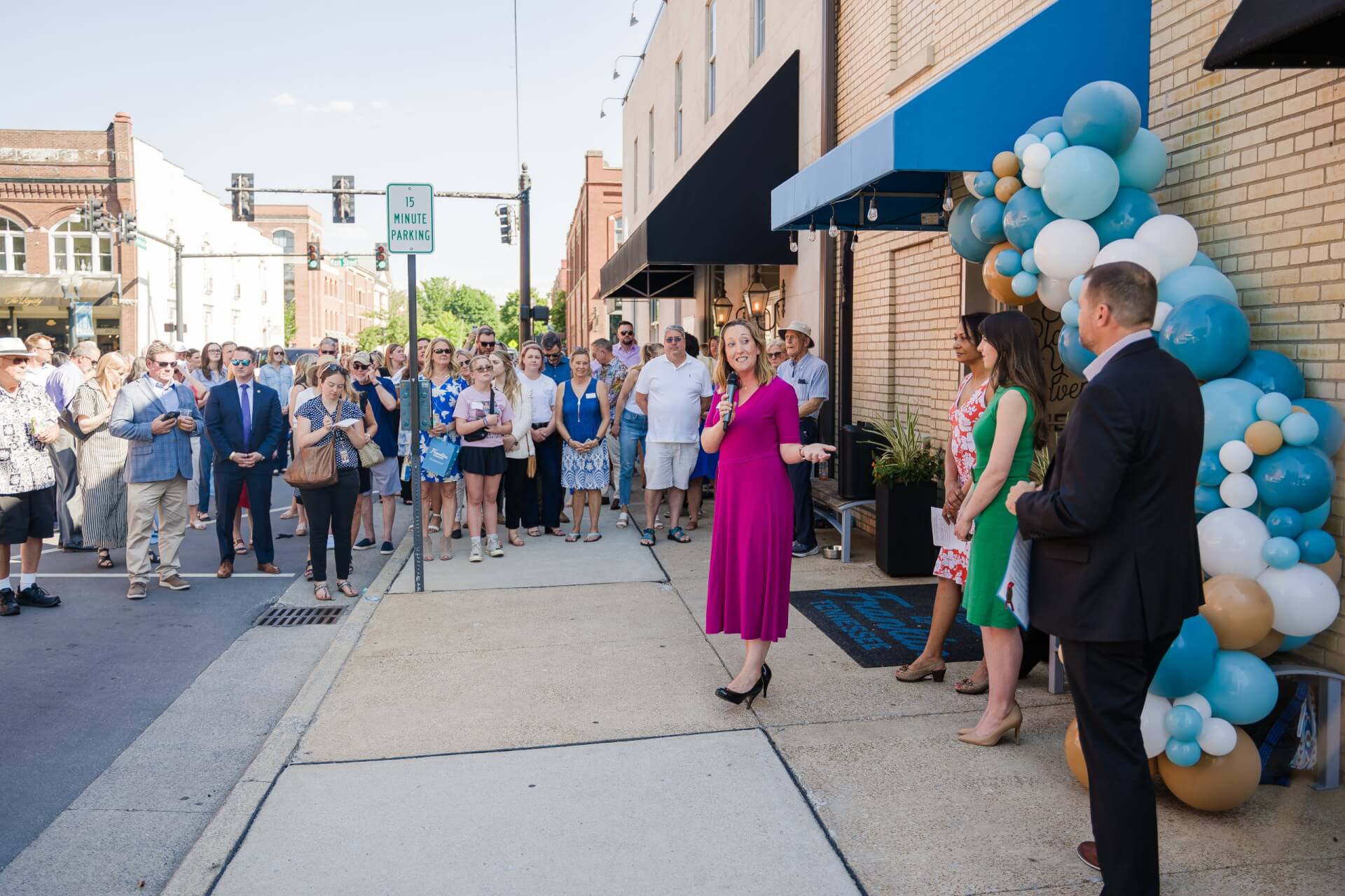 A vibrant street scene with a crowd gathered for a ribbon-cutting ceremony, featuring a woman in a pink dress speaking into a microphone, surrounded by colorful balloons and onlookers in Williamson County, Tennessee.