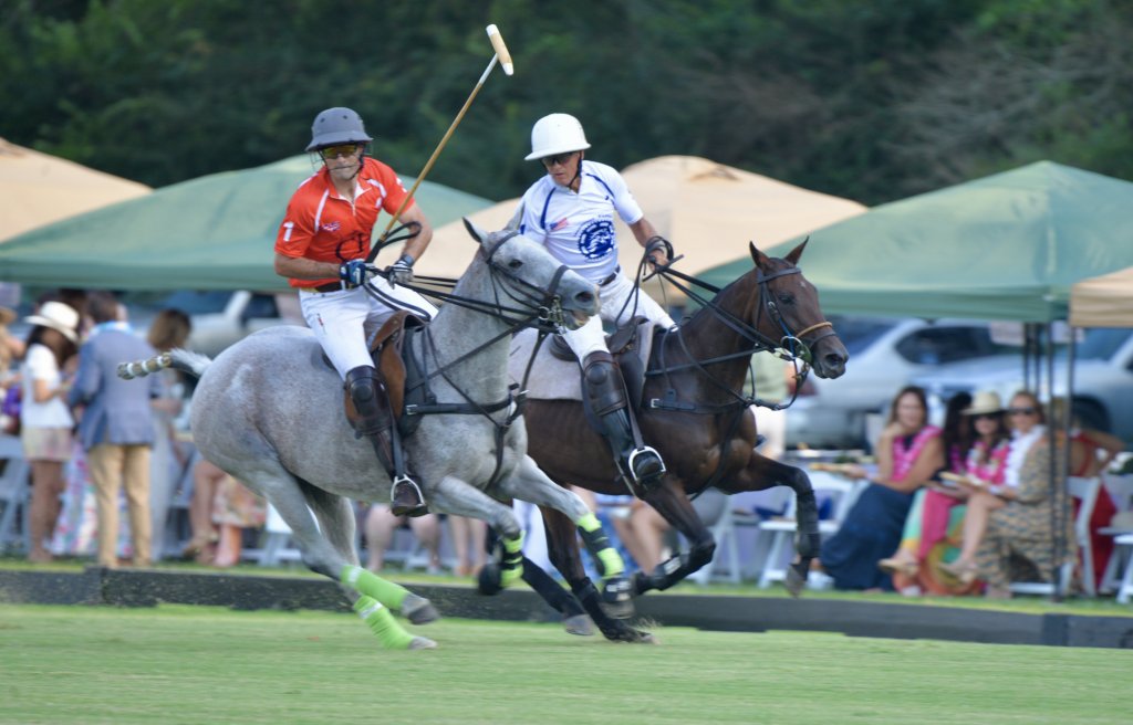 Two polo players on horseback, one in red and one in blue, compete in a match while spectators watch from under green tents in Williamson County, Tennessee.