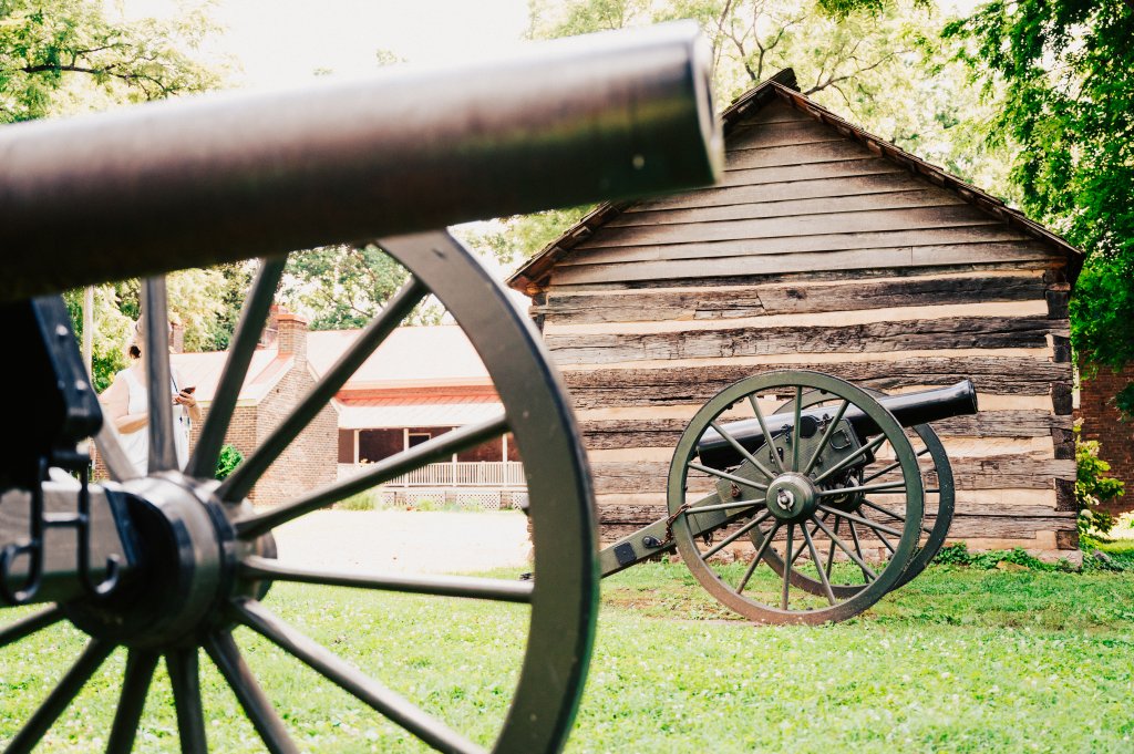 A cannon points towards a rustic log cabin surrounded by lush green grass and trees in Williamson County, Tennessee.