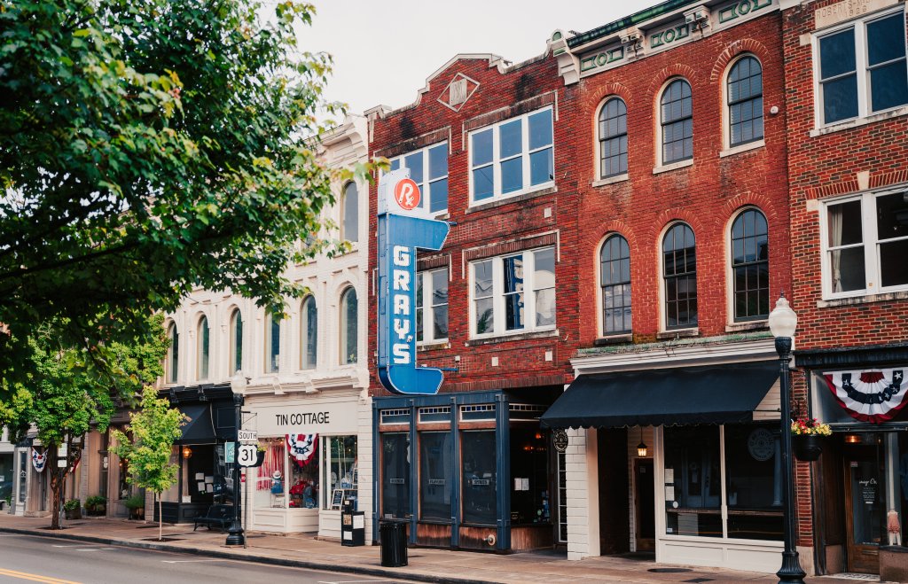 A charming downtown street lined with historic brick buildings, featuring a prominent blue neon sign for Gray's Drug Store and other quaint shops with awnings in Williamson County, Tennessee.