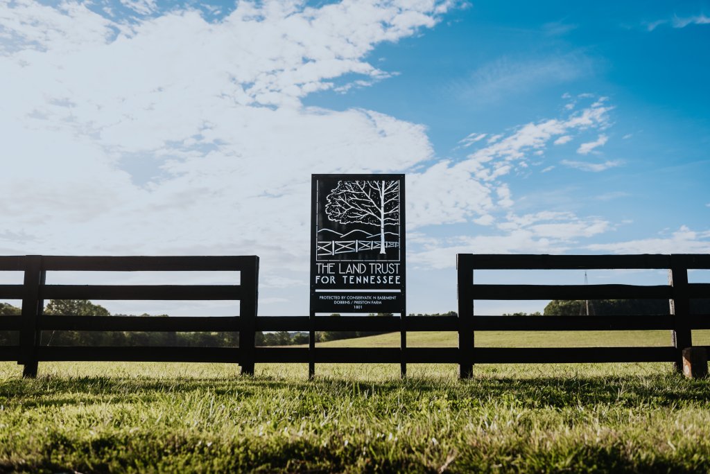 A wooden fence with a sign for The Land Trust for Tennessee in a grassy field under a blue sky with clouds in Williamson County, Tennessee.