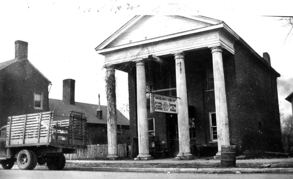 A vintage black and white photograph of a two-story brick building with four tall columns supporting a triangular pediment, situated on a street corner with a wooden truck parked in front and a residential house to the left in Williamson County, Tennessee.
