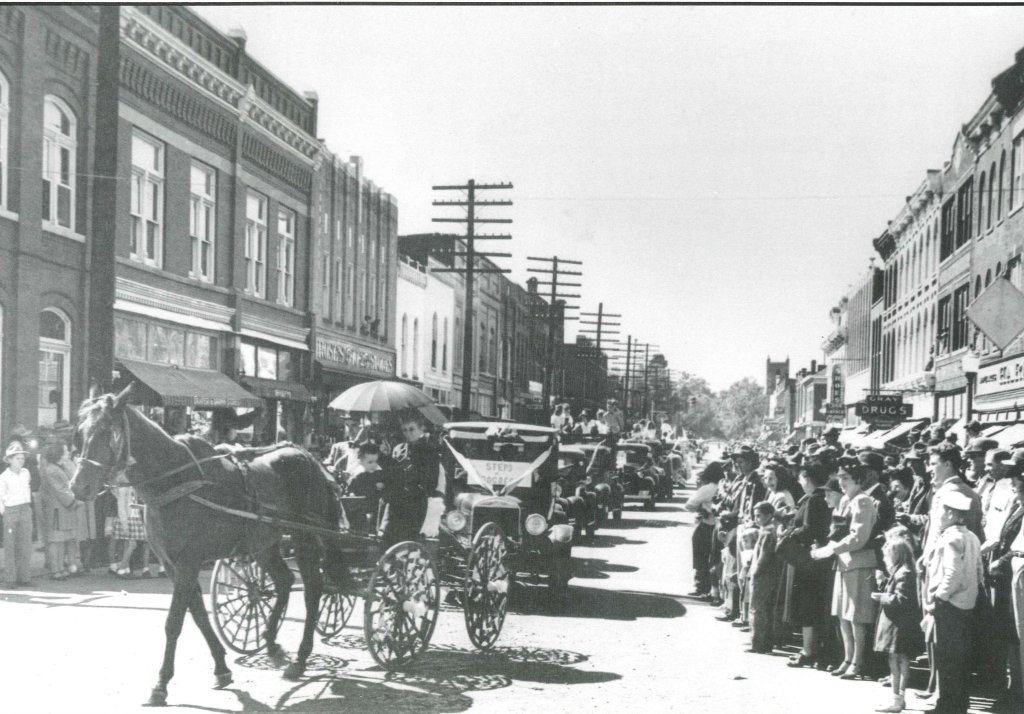 A bustling town square filled with people, horse-drawn carriages, and early 20th-century automobiles during a parade or celebration in Williamson County, Tennessee.