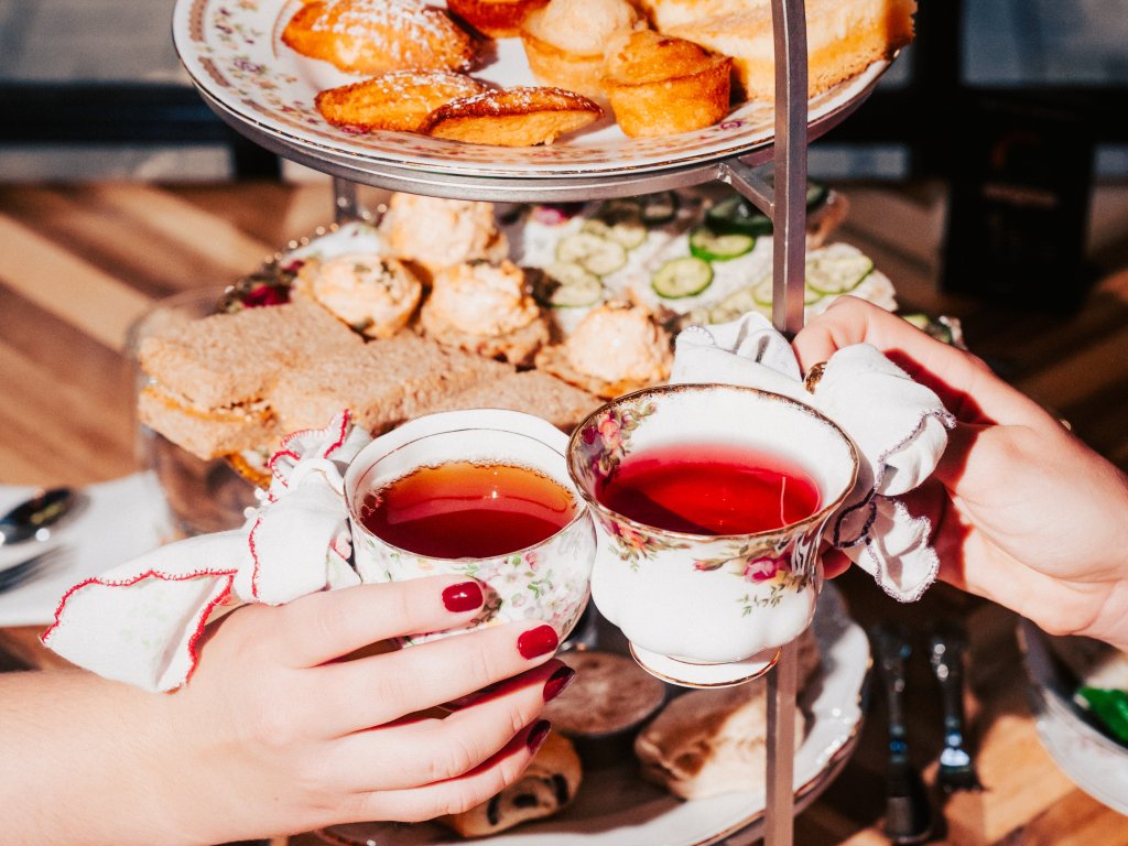 Two hands, adorned with red nail polish, gracefully clink delicate floral teacups filled with rich red tea, celebrating a moment of tea time indulgence amidst a lavish spread of scones, sandwiches, and pastries on a multi-tiered stand in Williamson County, Tennessee.