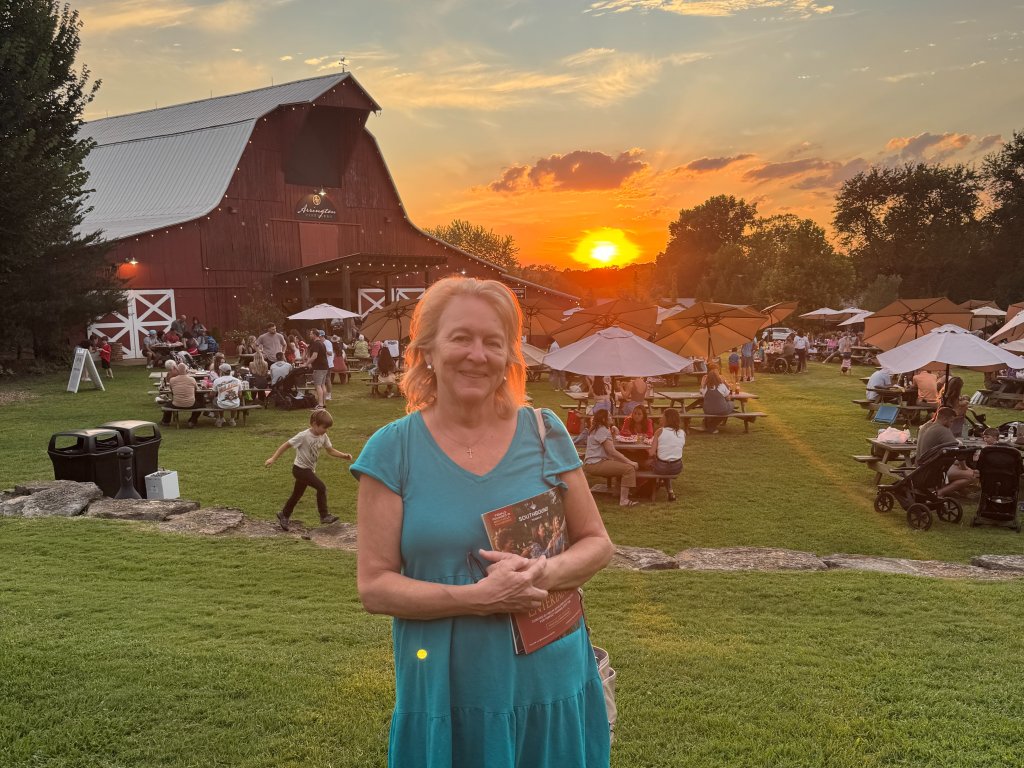 A woman in a blue dress stands smiling in a grassy field at sunset, holding a book, with a red barn and picnic tables in the background in Williamson County, Tennessee.