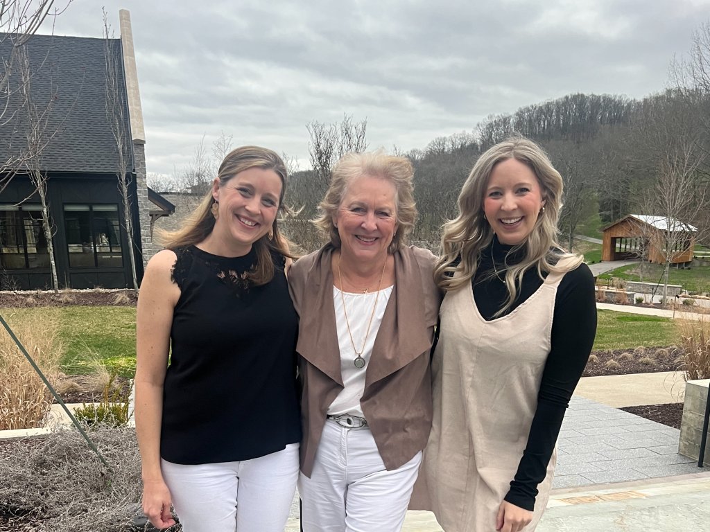 Three women stand together smiling in front of a modern house with a dark roof and large windows, surrounded by a landscaped garden with a wooden bridge in the background in Williamson County, Tennessee.