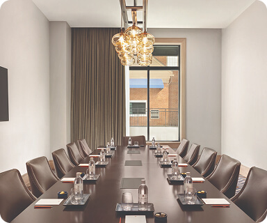 A modern conference room with a long wooden table, leather chairs, and a chandelier, featuring a large window with a view of a brick building and blue awning in Williamson County, Tennessee.