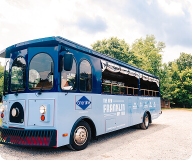 A blue trolley-style bus with 'The New Franklin' and 'Gray Line' branding parked on a gravel lot surrounded by trees in Williamson County, Tennessee.