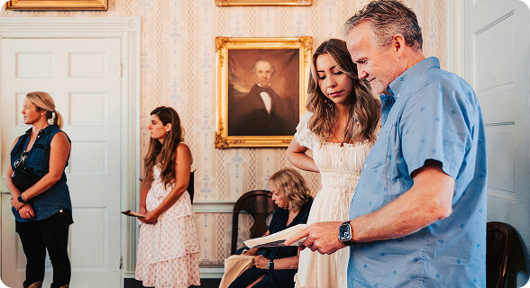 A group of people gathered in a room with a framed portrait on the wall, one man is reading a book to a woman in Williamson County, Tennessee.