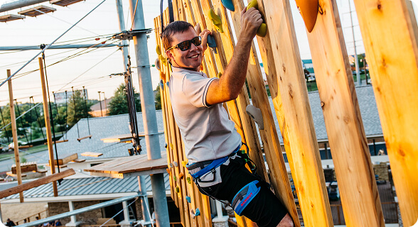 A man in a white t-shirt and sunglasses is rock climbing on a wooden wall with colorful handholds in Williamson County, Tennessee.