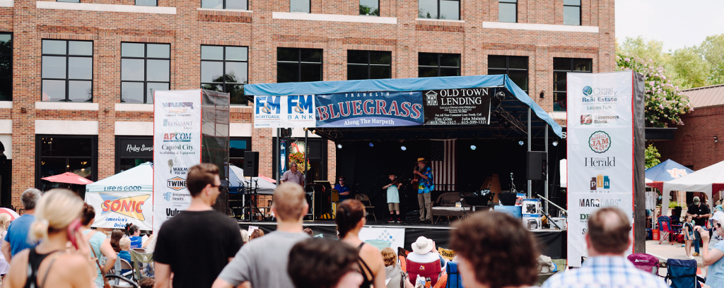Bluegrass Along the Harpeth A lively outdoor concert with a band performing on stage in front of a crowd, with banners and tents set up around the venue in Williamson County, Tennessee.
