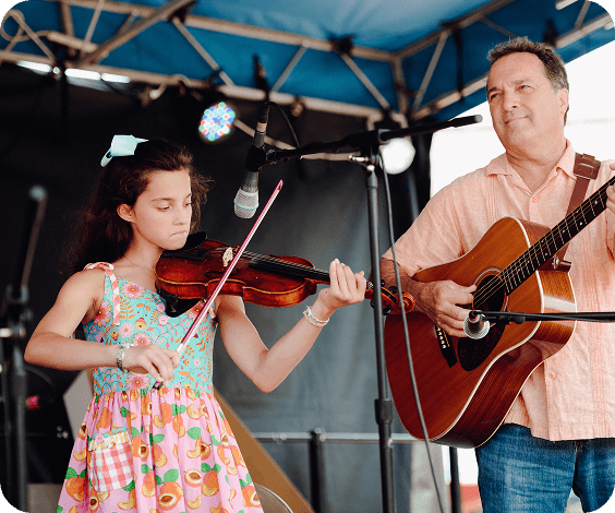 Bluegrass Along the Harpeth A young girl in a floral dress plays the violin while a man in a pink shirt accompanies her on an acoustic guitar on stage in Williamson County, Tennessee.
