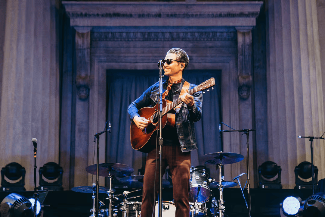 A musician performs on stage, playing an acoustic guitar and singing into a microphone, backed by a drum set and stage lights in Williamson County, Tennessee.