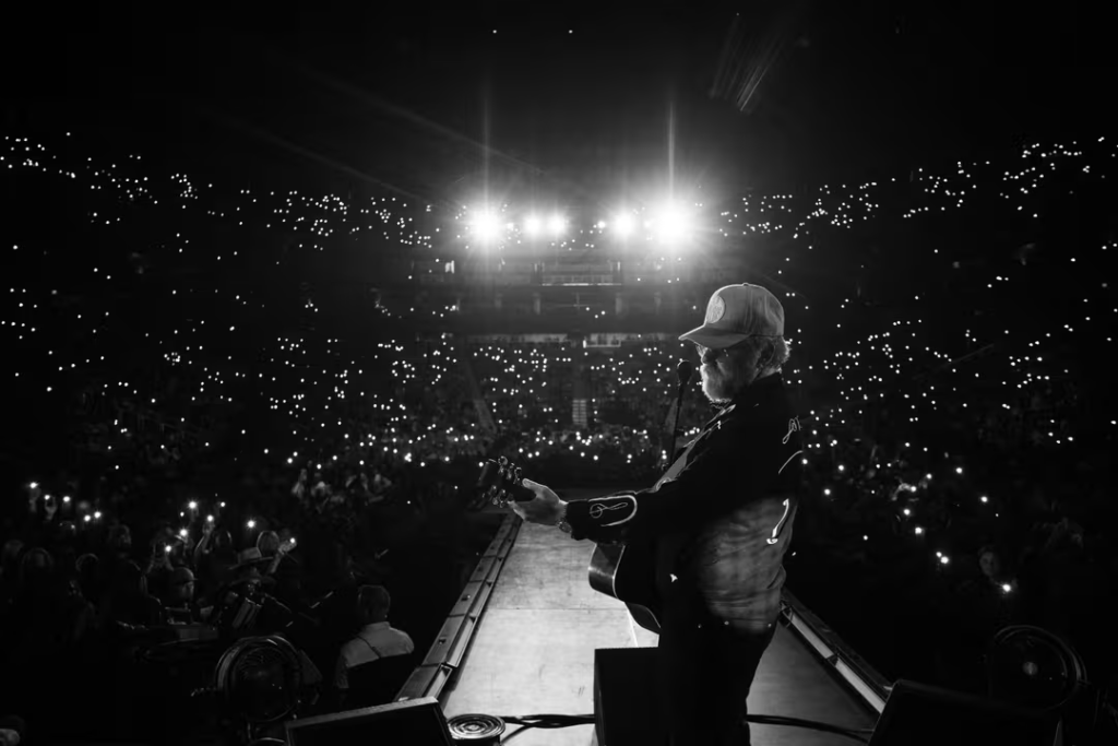 A musician performs on stage in a large arena filled with an enthusiastic crowd, illuminated by bright stage lights and a sea of glowing cell phones in Williamson County, Tennessee.