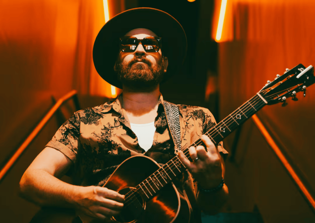 A man with a beard and sunglasses plays an acoustic guitar in a dimly lit room with red lighting in Williamson County, Tennessee.