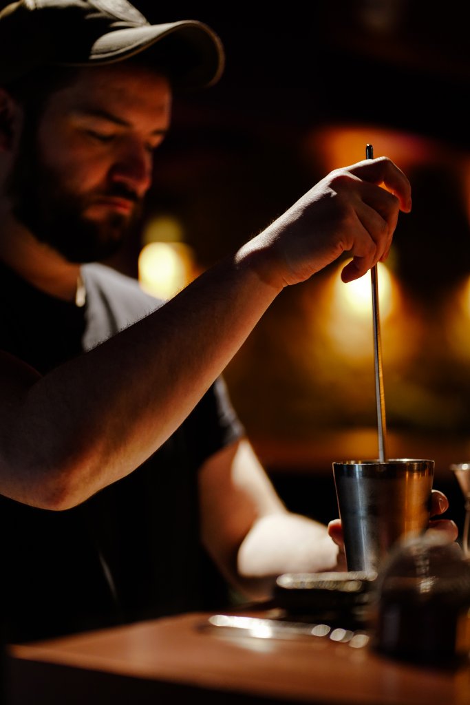 A bearded man in a dark setting, wearing a cap, carefully stirs a metallic cup with a long spoon in Williamson County, Tennessee.