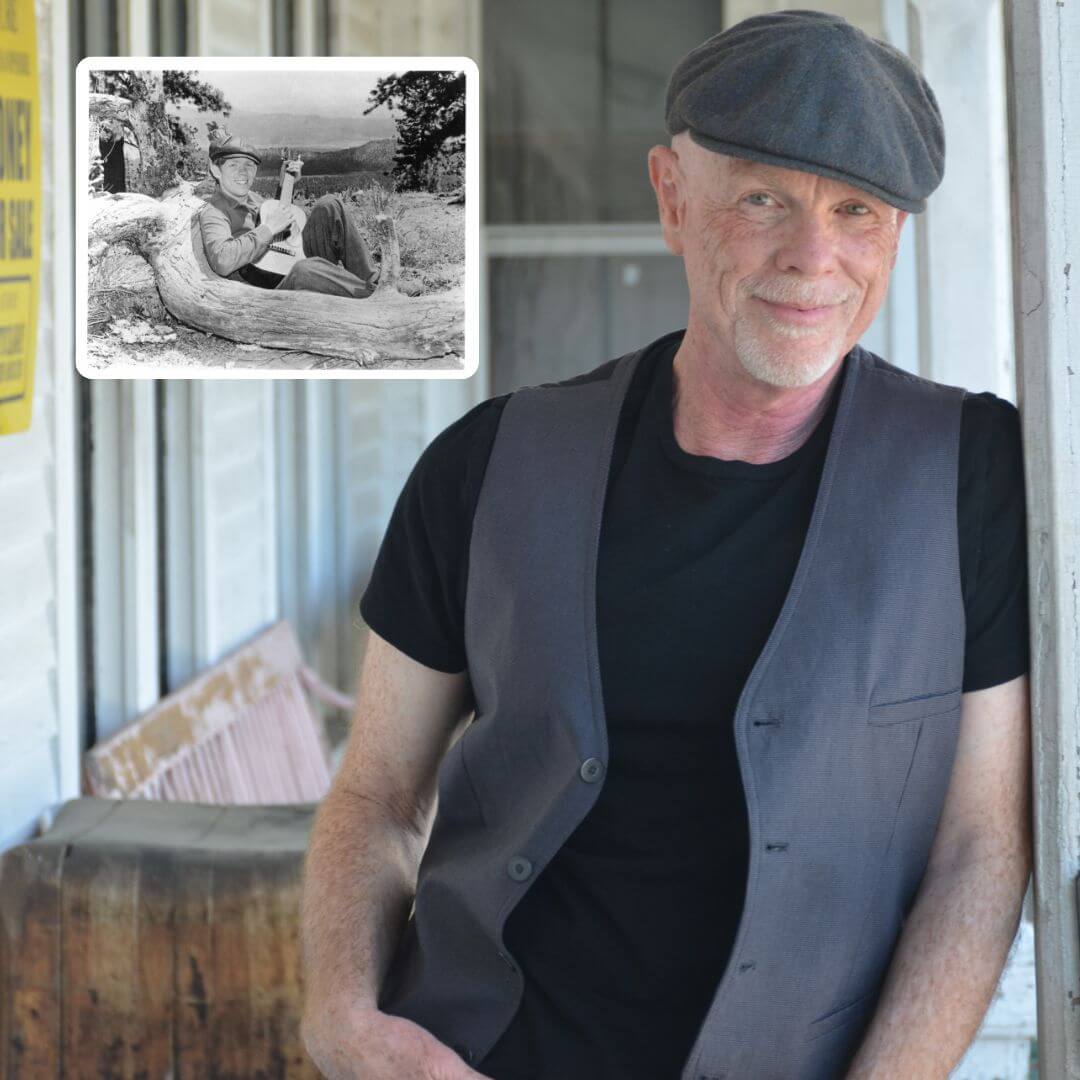 An elderly man with a bald head and a white beard, wearing a gray cap and a gray vest over a black shirt, stands smiling on a porch with a rustic wooden bench and a vintage black-and-white photograph of a man playing guitar on a log in the background in Williamson County, Tennessee.