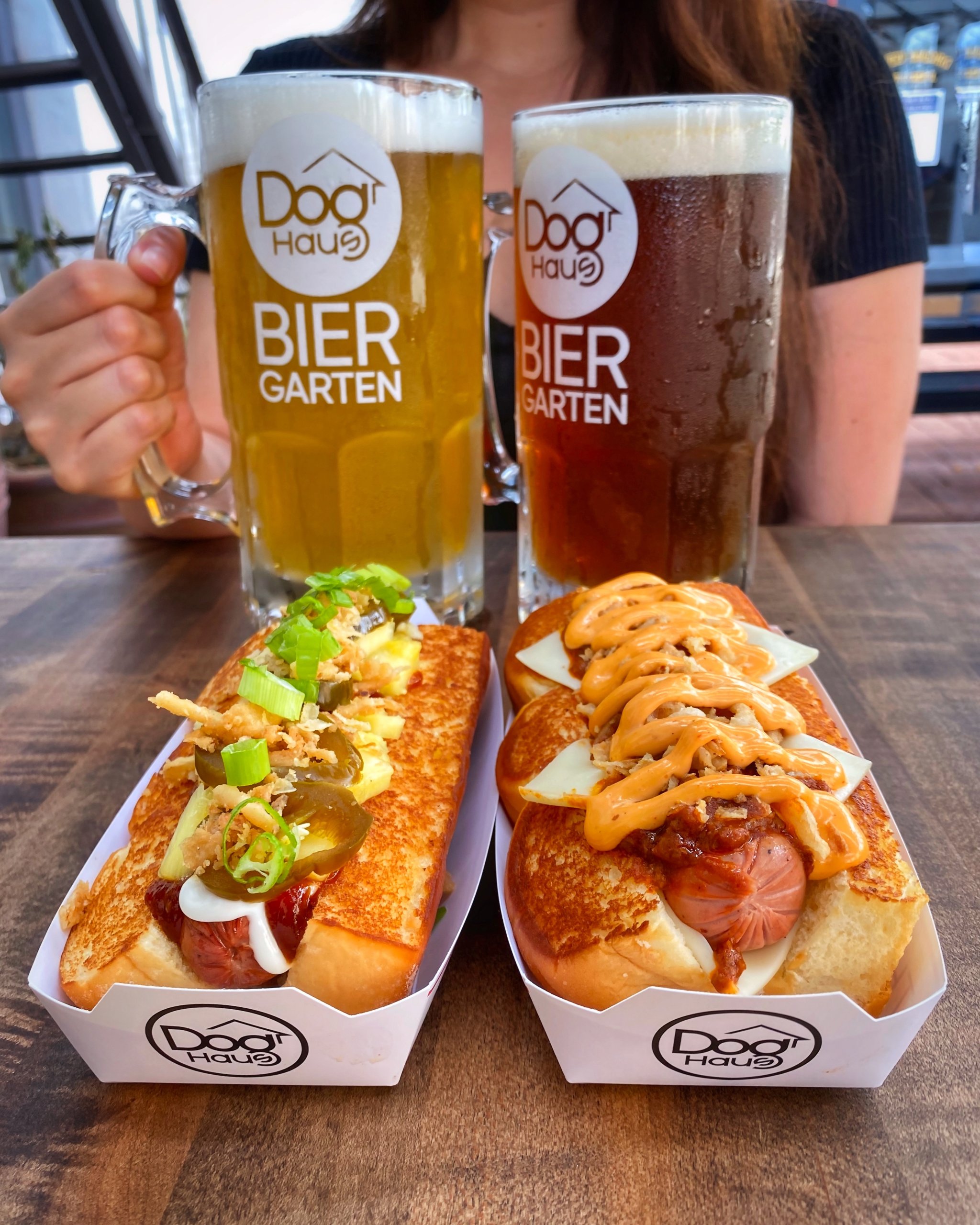 A woman holding two large beer steins labeled 'Doog Haus Bier Garten' with two gourmet hot dogs topped with various condiments in front of her on a wooden table in Williamson County, Tennessee.