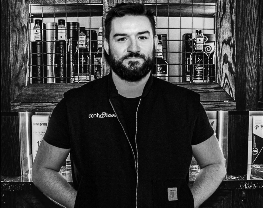 A man stands confidently in front of a well-stocked bar, wearing a black vest with 'onlydrama' embroidered on it in Williamson County, Tennessee.