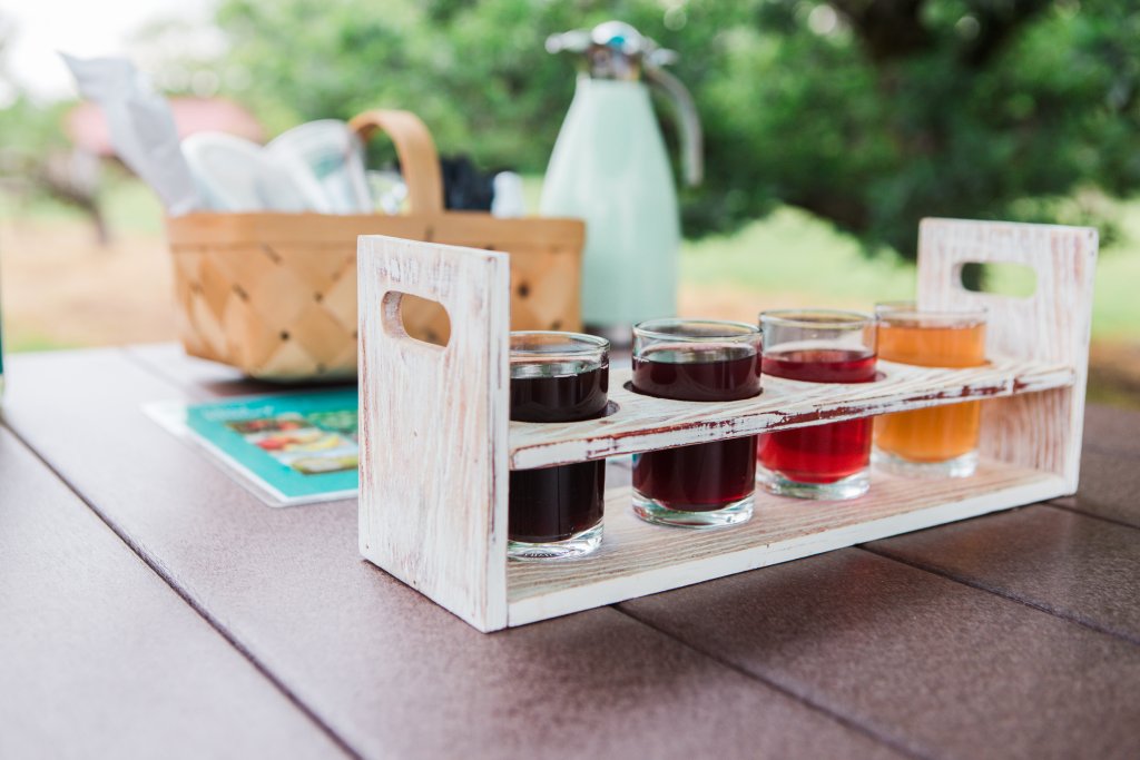 Events A wooden tray holds four glasses of colorful beverages, including red, dark red, and orange liquids, accompanied by a basket of utensils and a green pitcher on a wooden table outdoors in Williamson County, Tennessee.