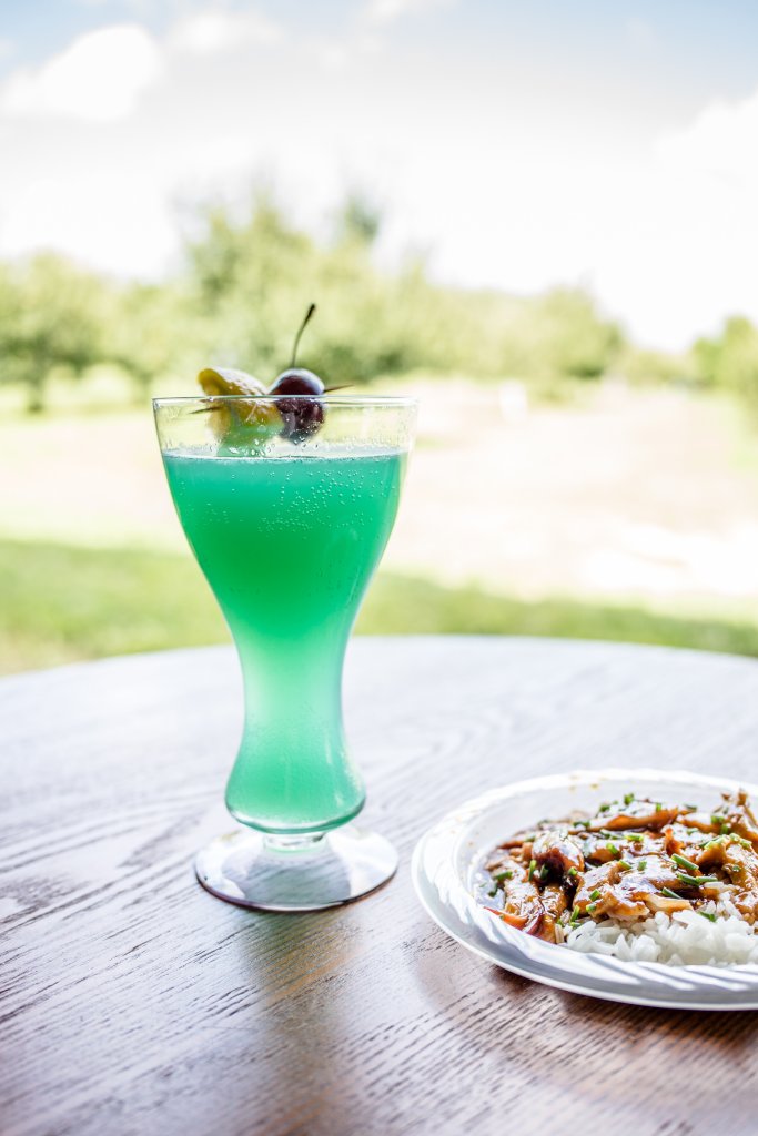 Events A vibrant turquoise cocktail garnished with a cherry and lemon slice sits beside a plate of stir-fried chicken and rice on a wooden table outdoors in Williamson County, Tennessee.