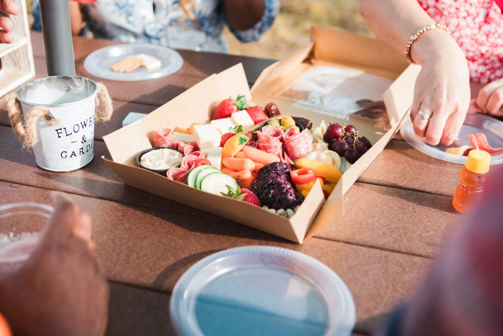 A colorful charcuterie board with fresh fruits, vegetables, meats, and cheeses is spread out on a wooden table, accompanied by a small bucket labeled 'Flower & Garden' and a bottle of honey in Williamson County, Tennessee.