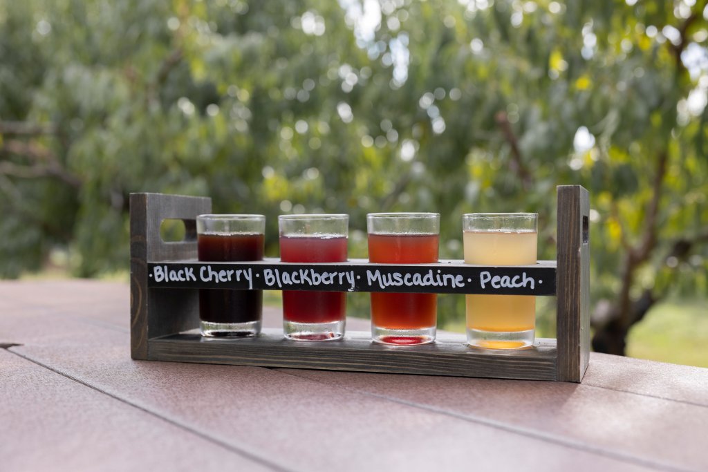 Events A wooden tray holds five glasses of different colored fruit juices, labeled as black cherry, blackberry, muscadine, and peach in Williamson County, Tennessee.