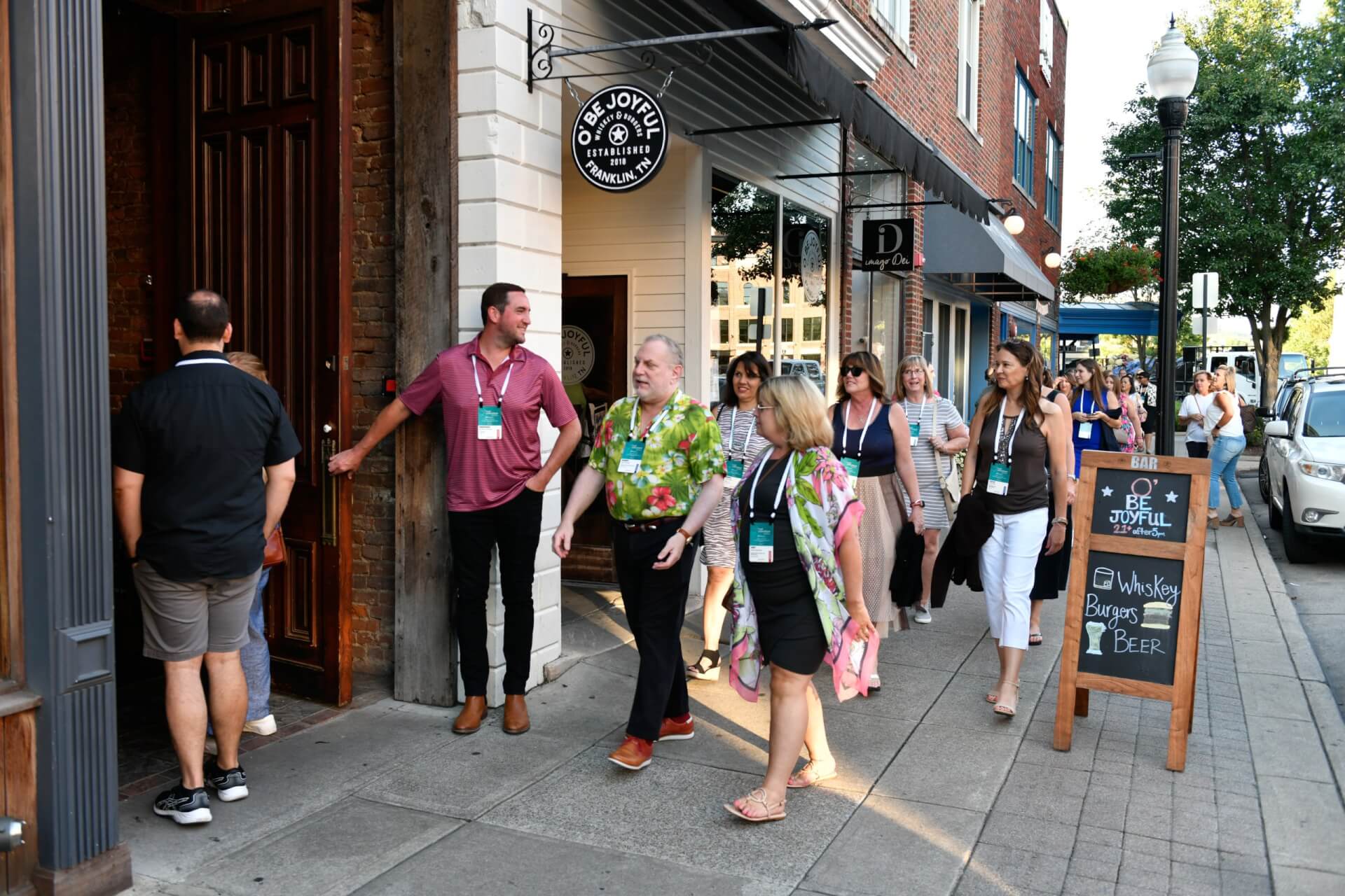 A group of people wearing conference badges walks along a brick sidewalk in front of a store with a sign reading 'Be Joyful' in Franklin, TN in Williamson County, Tennessee.